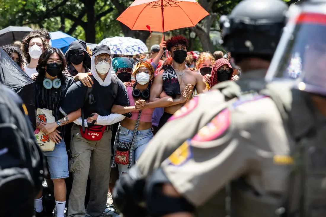 A line of protestors wearing N95s and other masks and carrying umbrellas stand with arms linked in front of police wearing riot gear.